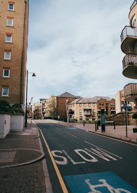 This image depicts a quiet urban street scene during daytime, with a downward-sloping road marked with a large white 'SLOW' sign on the asphalt and a designated blue accessible parking space adjacent to the curb. On the left side, there's a multi-story brick building with several small windows, while on the right, a modern residential building features several balconies with black railings. The street is lined with black bollards and a few pedestrians can be seen in the distance. A streetlight extends over the road, and the sky overhead is mostly cloudy with patches of blue. In the context of house removals and home relocation services, this setting could represent a typical route for furniture transport or packing and moving activities arranged by Man with Van Gunnersbury, demonstrating the urban environment where loading and unloading of boxes, furniture, and appliances may take place during a quick flat-move between Acton Lane and Gunnersbury.