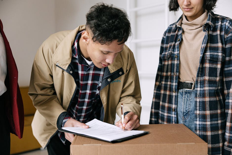 A man dressed in a beige jacket and a plaid shirt is signing a document placed on top of a cardboard box, likely during a home relocation or moving process. He is inside a property, near a doorway or entry point, and is focused on the paperwork, which has a pen in his right hand. Standing next to him is a woman wearing a checkered shirt over a beige top, observing the signing. In the background, there are visible shelves and a neutral coloured wall, with soft lighting illuminating the scene. The cardboard box in question may contain household items or packed belongings ready for transport. The setting indicates a preparatory stage of furniture transport or packing during a house move, with the involvement of professional removals services by Man with Van Gunnersbury, supporting efficient logistics for the move from Acton Lane to Gunnersbury.