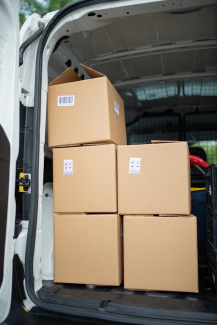 Inside the rear of a white van used for home relocation, four large cardboard boxes are stacked neatly, with two on the floor and two placed on top. Each box has a white barcode label affixed to its side, indicating organized packing. The boxes are made of brown corrugated cardboard, and the interior of the van features a metal ceiling with a reflective surface. To the right, a black shopping trolley with a red handle is partially visible, used for transporting smaller items or packing materials. The van is parked on a pavement near a property, suggesting an active loading process typical of furniture transport during a house removal. The lighting inside the van is natural, coming through the open door, and the scene captures a professional approach to packing and loading, consistent with services offered by Man with Van Gunnersbury.
