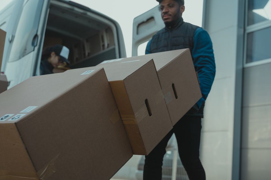 A man wearing a dark blue jacket and black trousers is standing outside a white moving van, carrying three medium-sized cardboard boxes stacked one on top of the other, secured with packing tape. In the background, another person is partially visible inside the van, leaning over and handling additional packing materials or boxes. The scene is set on a paved area adjacent to a building with a closed roller shutter, and a white door is visible to the right. The van’s sliding door is open, revealing the interior with more boxes and packing supplies, indicative of a home relocation or furniture transport process, as managed by Man with Van Gunnersbury. The lighting suggests daytime, with natural light illuminating the scene, emphasizing the careful handling of packing materials during the loading process involved in house removals.