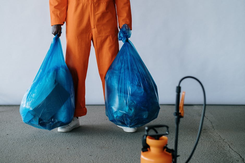 A person dressed in orange work overalls and white footwear is standing indoors on a concrete floor, holding two large blue plastic waste bags filled with rubbish. The individual’s torso and legs are visible, but their face is not shown. In the foreground, there is an orange handheld sprayer attached to a hose, and a black and orange pump spray bottle placed on the floor. The background features a plain light grey wall, creating a neutral setting suitable for describing home relocation or waste removal services. The scene captures the process of clearing bulky waste items for disposal or removal during a house move, illustrating the handling and transport of waste materials as part of a professional removals or clearance service by Man with Van Gunnersbury.