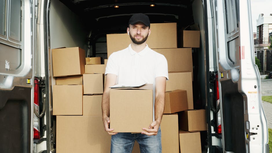 A man wearing a white polo shirt and a black cap stands inside the open rear of a white delivery van, which is loaded with numerous stacked cardboard boxes, some of which are of small size and others larger, secured with packing materials. The man is holding a cardboard box in front of him, with some paperwork resting on top of it. The van's interior appears to be dedicated to furniture transport and packing, with the boxes neatly arranged on a flat surface. Outside, a suburban residential area with houses, greenery, and a pavement is visible, indicating the loading process in a home relocation or house moving scenario. The lighting is natural, suggesting daytime, and the environment shows signs of an organized move, possibly facilitated by Man with Van Gunnersbury, a professional removals service specializing in house removals and furniture transport.