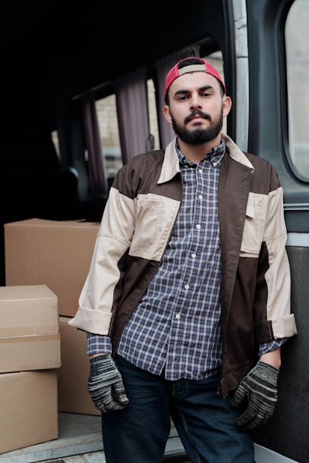 A man with dark hair and a beard, wearing a red cap, casual checkered shirt, and a tan and brown jacket, stands inside the back of a van used for house removals during a furniture transport process. The van's interior features visible dark upholstery and a sliding door open to reveal the loading space, which contains neatly stacked cardboard boxes of various sizes wrapped in brown packaging tape. The man is wearing grey work gloves and has his hands relaxed at his sides, preparing for or completing a transfer of belongings. Natural light from outside illuminates the interior, highlighting the surrounding objects and the vehicle's interior. This scene exemplifies home relocation logistics managed by Man with Van Gunnersbury, illustrating the packing and loading aspects of a professional removals service supporting furniture transport and house moves.