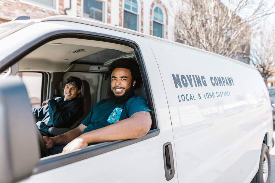 Inside the cab of a white moving van with the sliding door open, two movers are visible, sitting in the driver and passenger seats. The mover in the driver's seat is a man with dark, curly hair and a 