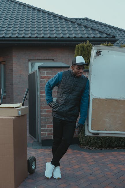 Inside the cab of a white moving van with the sliding door open, two movers are visible, sitting in the driver and passenger seats. The mover in the driver's seat is a man with dark, curly hair and a 