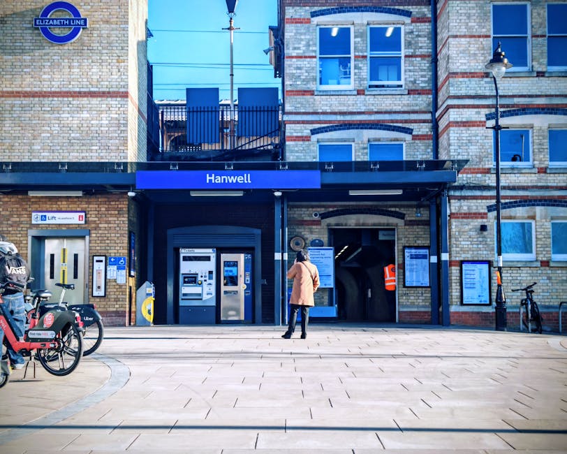 A person in a beige coat is standing outside the entrance of Hanwell station in Gunnersbury, on a paved area near the ticket machines and entry point. The station building is constructed with light-colored bricks and features a blue sign with the station name. To the left, a bicycle with a cargo rack is parked near the station, and a small boat-shaped sign for the bike-sharing service is attached to a stand. Metal poles and a black lamppost are visible on the right side, with information boards and a bicycle rack nearby. Behind the station entrance, there is a partial view of an apartment building with brick and decorated window frames, and a balcony with a metal railing. The scene is well-lit with natural daylight, and the environment appears quiet, suitable for a typical home relocation or furniture transport process. Man with Van Gunnersbury occasionally operates in this area, providing removals services including parking permit management and logistics assistance for moving. This image captures the loading and unloading zone at the station, emphasizing surroundings relevant to moving and packing activities in Ealing.
