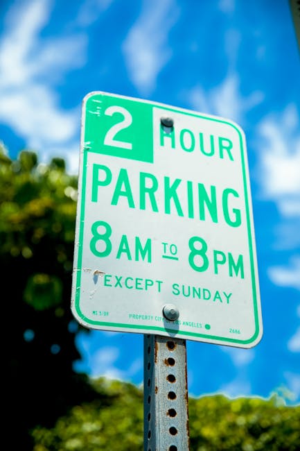 A green and white parking restriction sign mounted on a metal post outdoors, indicating a two-hour parking limit from 8 a.m. to 8 p.m., Monday to Saturday, with exceptions on Sundays. The sign is set against a bright blue sky with some white clouds and green leafy trees in the background. The sign's text is clear and legible, with the top portion showing a bold '2' and the words 'HOUR PARKING' beneath, followed by the time details and the note 'EXCEPT SUNDAY.' This type of signage is relevant to moving and removal services, such as those provided by Man with Van Gunnersbury, when planning home relocations and furniture transport in busy urban areas, ensuring compliance with local parking regulations during loading and unloading processes.