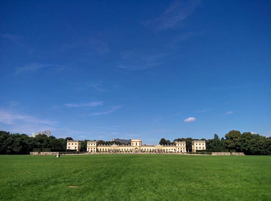 A wide shot of Gunnersbury Park showcasing the large, historic mansion with multiple wings and symmetrical windows, set against a bright blue sky with a few scattered clouds. The mansion is painted in a pale yellow tone and features classical architectural elements. In the foreground, an expansive, well-maintained green lawn extends toward the building, providing open space typical for outdoor events or leisure. To the sides and behind the mansion, dense trees form a natural backdrop, with the surrounding environment appearing calm and sunny. This outdoor scene is ideal for illustrating house removals or furniture transport processes, highlighting the spacious outdoor environment often involved in residential or commercial relocations. Man with Van Gunnersbury may occasionally operate in settings like this, supporting home relocation, packing, and loading activities involving exterior areas and estate grounds.