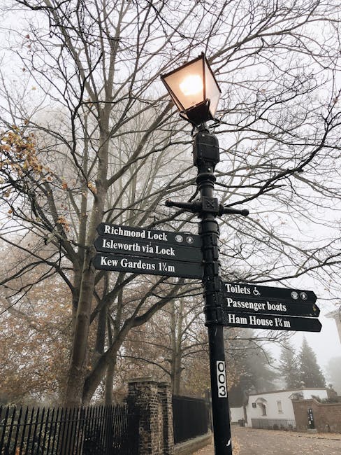 A black metal street lamp mounted on a tall post with a lit lantern at the top, situated outdoors amidst leafless tree branches on a foggy day. Attached to the post are multiple black directional signage boards with white text, indicating local destinations such as Richmond Lock, Isleworth via Lock, Kew Gardens 1/4 mile, Toilets and Passenger Boats, and Ham House 1/4 mile. The background features a partly visible brick wall, a small white structure, and overcast sky, reflecting a typical scene encountered during home relocation or moving processes in the Gunnersbury area, with signage supporting navigation to nearby historic landmarks and amenities.