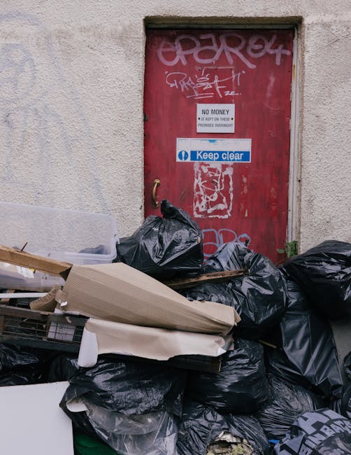 An outdoor scene showing a red metal door covered in graffiti, attached to a beige textured wall. Several black garbage bags and packaging materials, including cardboard and plastic wrapping, are piled on the pavement in front of the door. Some of the bags appear full and tightly sealed, with discarded packaging and furniture wrapping visible among the debris. The area suggests waste or unwanted items awaiting collection, with a small green plant growing near the base of the building. This setting relates to waste disposal often encountered during home relocation or moving processes. Occasionally, Man with Van Gunnersbury handles such waste removal as part of comprehensive house removals and packing services, ensuring clean and clear premises for efficient furniture transport and moving activities.