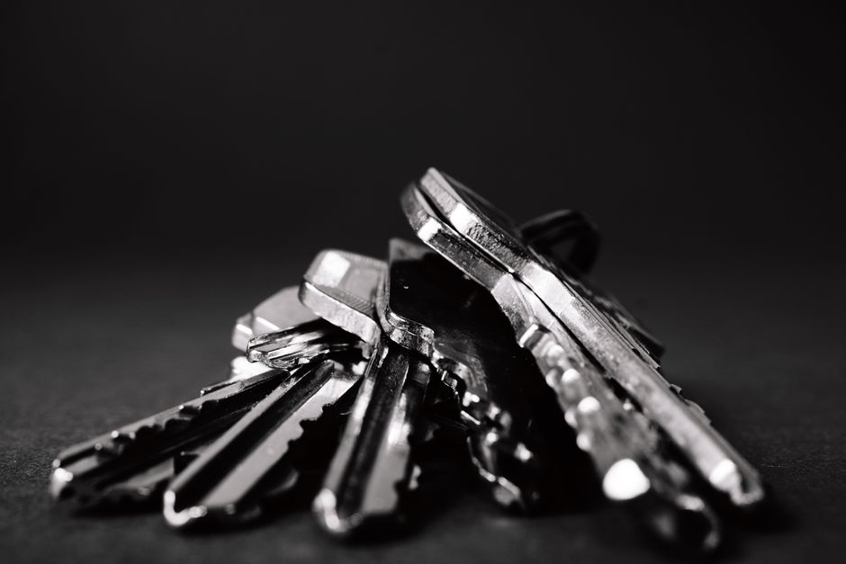 A close-up image showing one person handing over a set of house keys attached to a keyring to another individual. The person receiving the keys has an open palm positioned horizontally, with a light-colored wristwatch visible on their wrist, and is wearing a white wristband. The person giving the keys has a darker skin tone, is wearing a black wristwatch, and is holding the keys with their fingers, which are visible in the image. The background features a light green wall, suggesting an indoor setting, possibly within a property involved in a home relocation or moving process. The scene captures a moment of key exchange during the moving or furniture transport phase, associated with professional removals services such as those offered by Man with Van Gunnersbury.