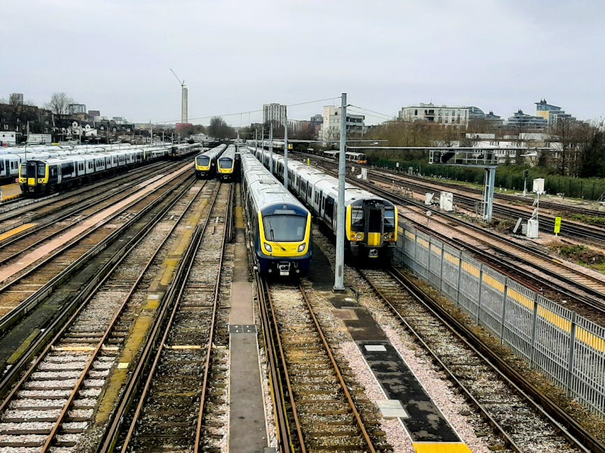 A collection of modern trains parked on multiple railway tracks adjacent to a station platform in an urban setting. The trains feature sleek, streamlined designs with yellow and grey liveries, and are positioned with some facing forward and others alongside each other. Overhead electric lines run above the tracks, supported by metal poles, indicating electric train services. In the background, there are residential and commercial buildings, including mid-rise apartments and office blocks, with some greenery visible in the distance. The platform area appears clear, with no passengers present, and the scene is captured during daylight under an overcast sky. This setting relates to train and rail transport, exemplifying the type of infrastructure involved in home relocation or moving logistics if using railway freight or services like those provided by Man with Van Gunnersbury.