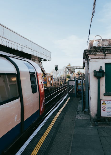 A view of a train arriving at Gunnersbury Station platform, with a white and red electric train on the left side of the image. The platform is paved with tactile paving strips along the edge for accessibility, and a small white building with a green window shutter is positioned on the right side, featuring signage and a black mailbox attached to its wall. Overhead, electrical lines are visible against a partly cloudy sky. In the background, railway tracks extend into the distance, with some freight containers and station infrastructure visible. This scene illustrates a typical urban railway station environment used for home relocation plans involving furniture transport, packing, and moving logistics. The presence of the station surroundings and the train emphasizes the importance of efficient transport routes for local removals services, such as those offered by Man with Van Gunnersbury.