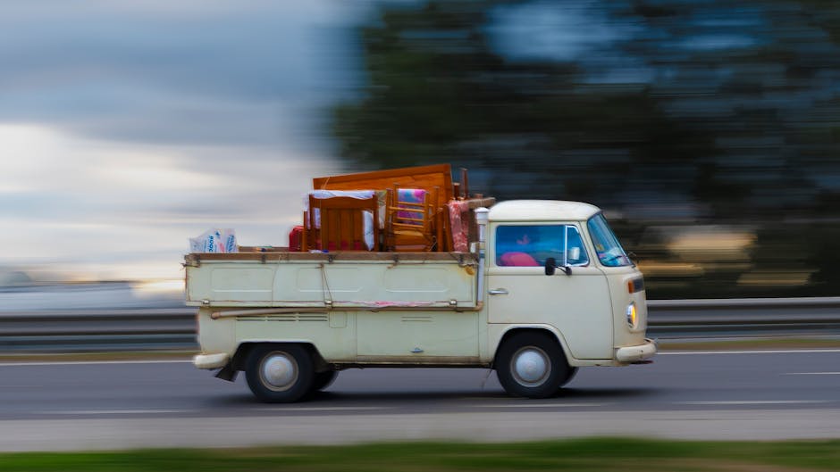 A small vintage off-white van with a boxy shape is moving along a roadway during daytime, with a blurred background indicating motion. The cargo area at the back of the van is loaded with various hous